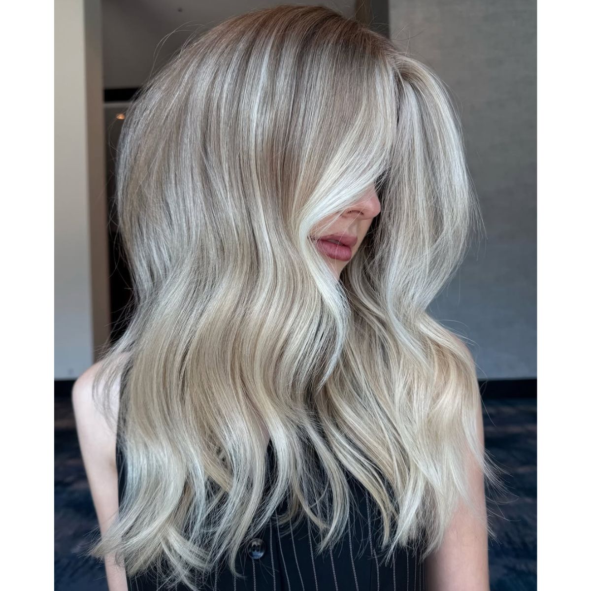 A woman with voluminous, wavy platinum blonde hair and bright, cool-toned highlights, styled with a deep side part and smooth, glossy texture in the same indoor location.
