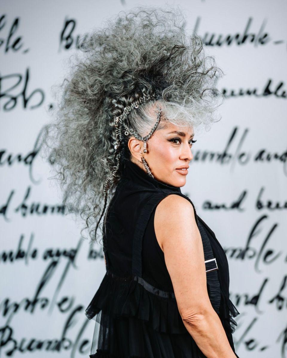 A model poses in front of a Bumble and bumble backdrop with a voluminous gray curly updo, decorative hair accessories and statement earrings.