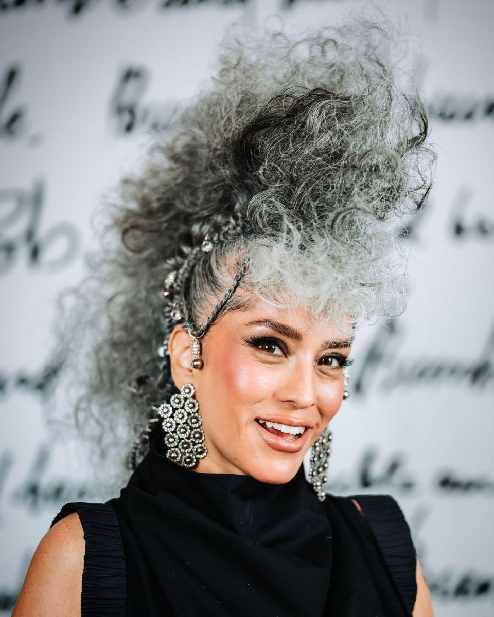 A model poses in front of a Bumble and bumble backdrop with a voluminous gray curly updo, decorative hair accessories and statement earrings.