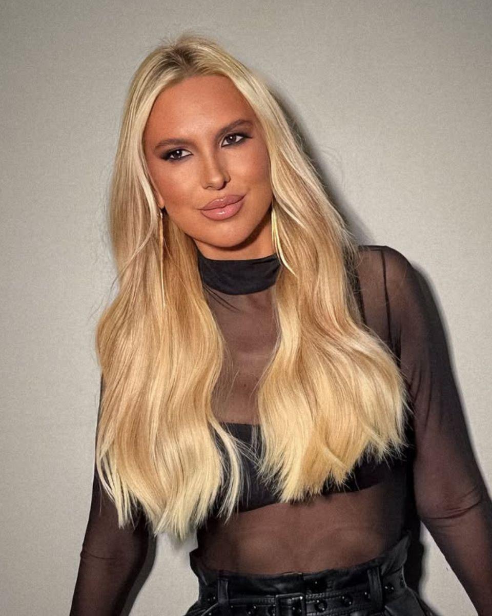 Portrait of long champagne blonde hair with soft beige dimension, a seamless root blend and polished waves styled over a sheer black top.