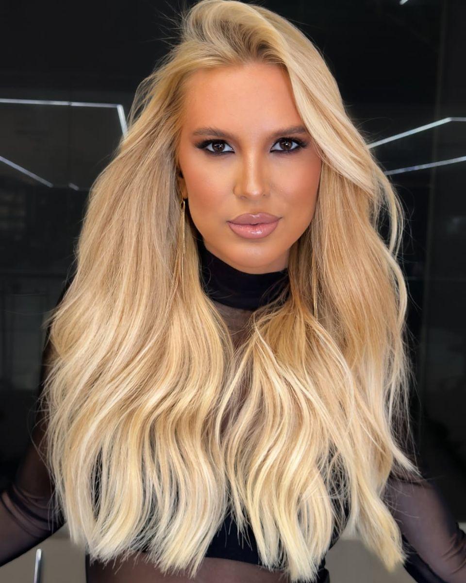 Portrait of long champagne blonde hair with soft beige dimension, a seamless root blend and polished waves styled over a sheer black top.