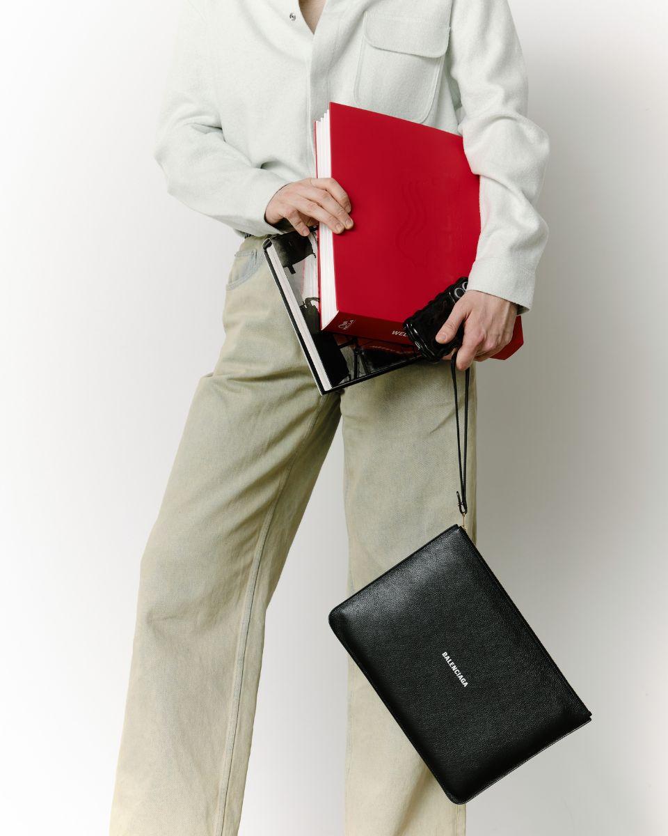 Chris Weber holds a red Wella portfolio and black Balenciaga bag while wearing a light shirt and neutral pants.