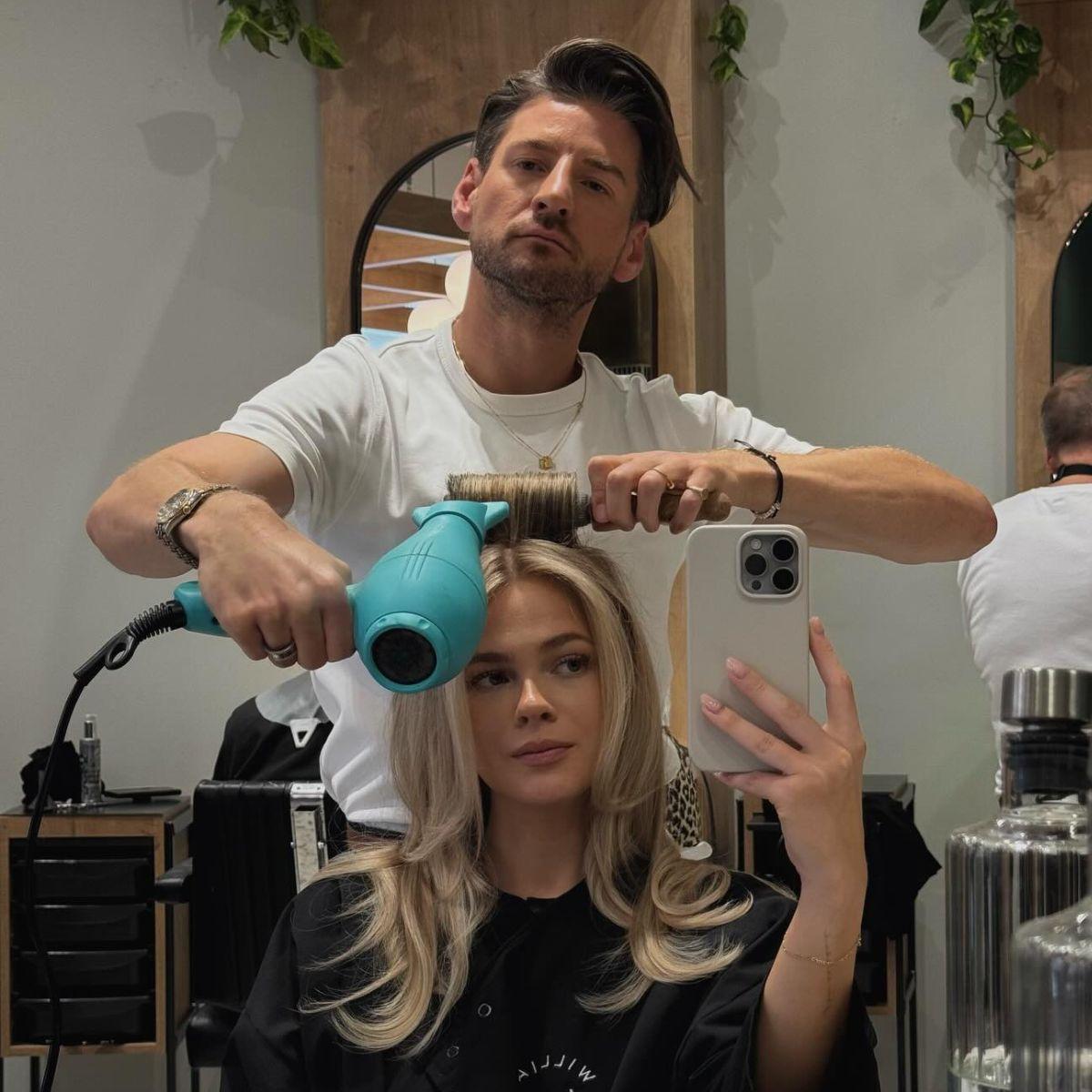 A hairstylist blow-dries a client’s freshly colored blonde hair in a salon. The client, seated in front of a mirror, takes a selfie while the stylist styles her voluminous, layered blonde look.