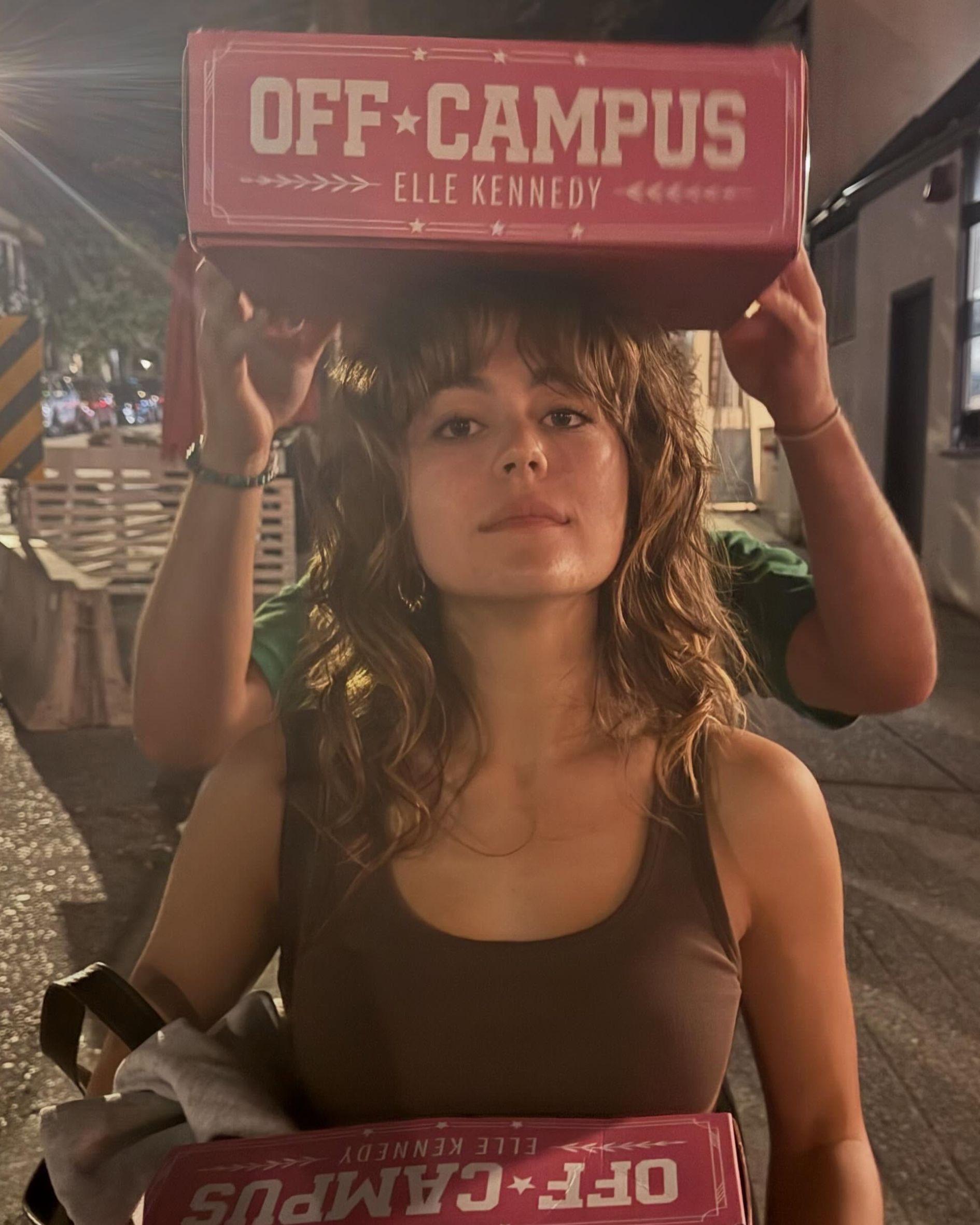 Behind-the-scenes photo of Mika Abdalla balancing oversized Off Campus book props on her head during the Off Campus press tour. She wears a brown tank top and her signature shoulder-length curly shag haircut with wispy bangs, standing outdoors at night.