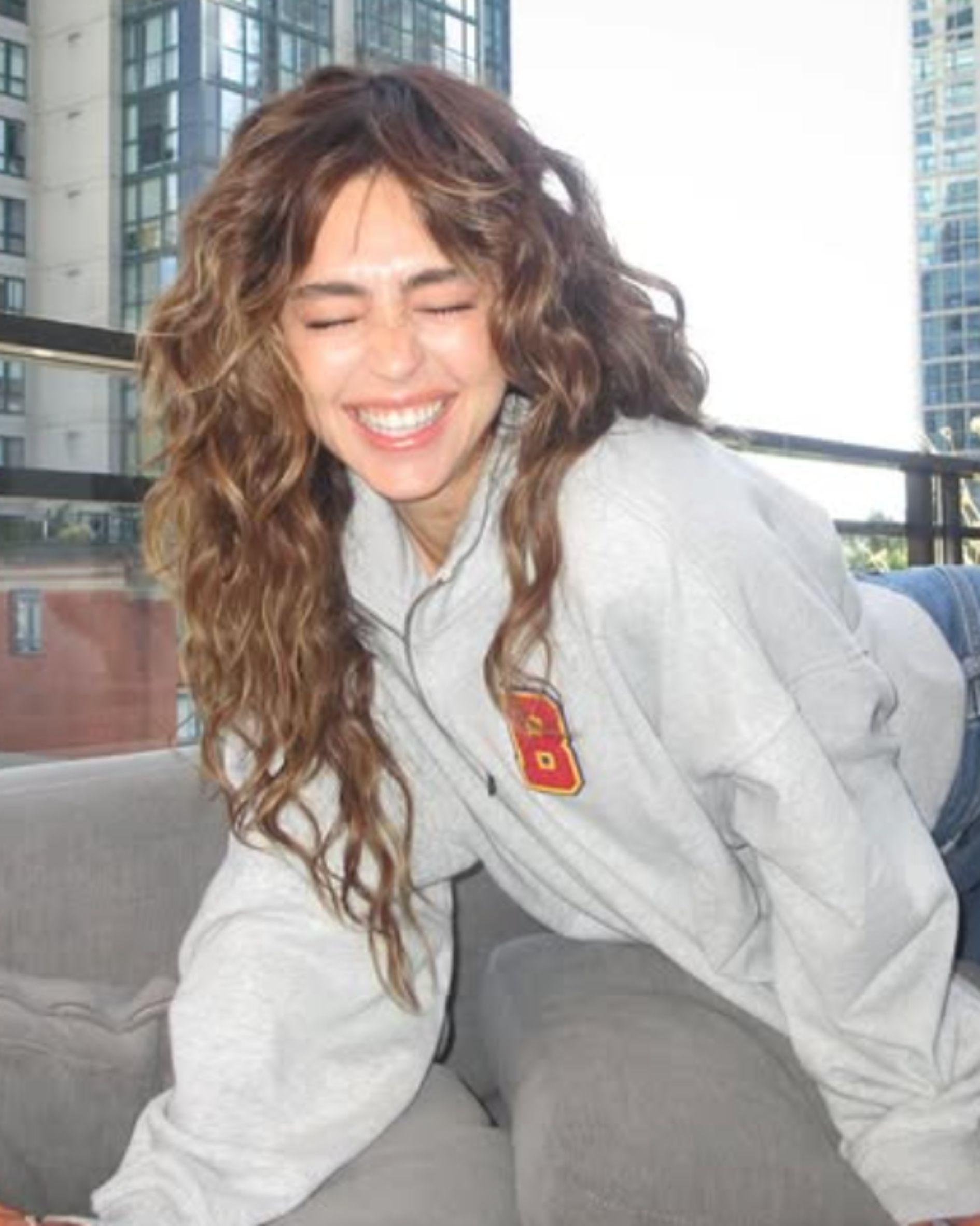 Mika Abdalla smiles with her eyes closed while showing off her long, textured curly hairstyle with face-framing layers and wispy curtain bangs. She wears a gray collegiate sweatshirt and is photographed on a city balcony during the Off Campus press tour.