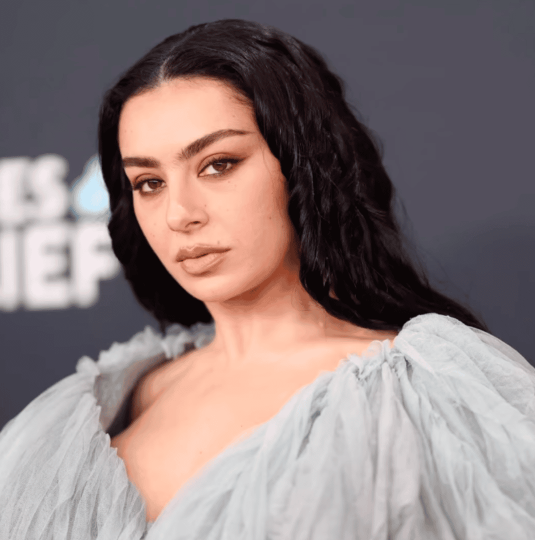 Charlie XCX posing at a red carpet event in a sheer light blue gown with wavy black hair.