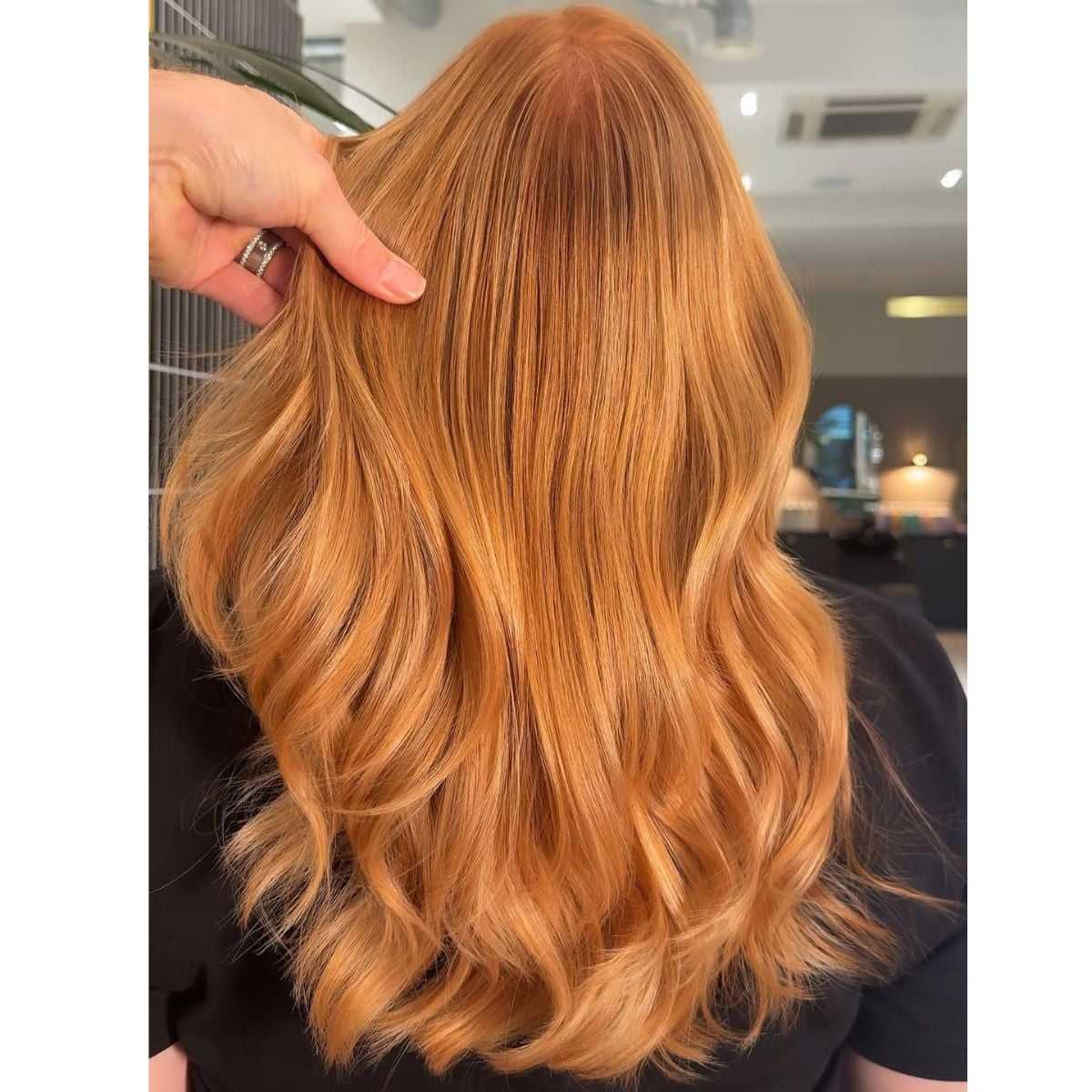 A person with long, wavy copper hair featuring a rich, golden glow. The hair is styled in loose waves, and a hand is gently lifting a section to show the color’s depth and shine in a salon setting.