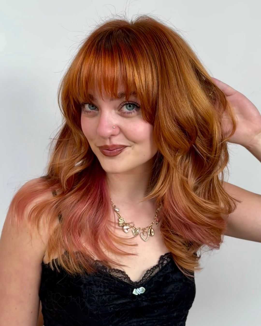 Front-facing portrait of a model wearing a dimensional ginger copper hair color with curtain-style fringe, soft curls and pastel pink accents blended through the ends.