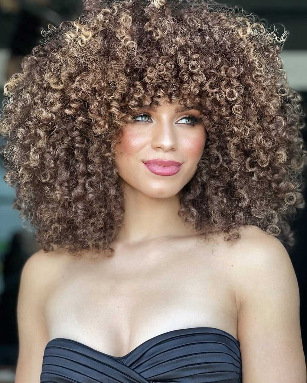 Side-angle view of voluminous curly hair featuring a dimensional mocha bronde color with beige-blonde ribbon highlights that enhance curl definition and shine.