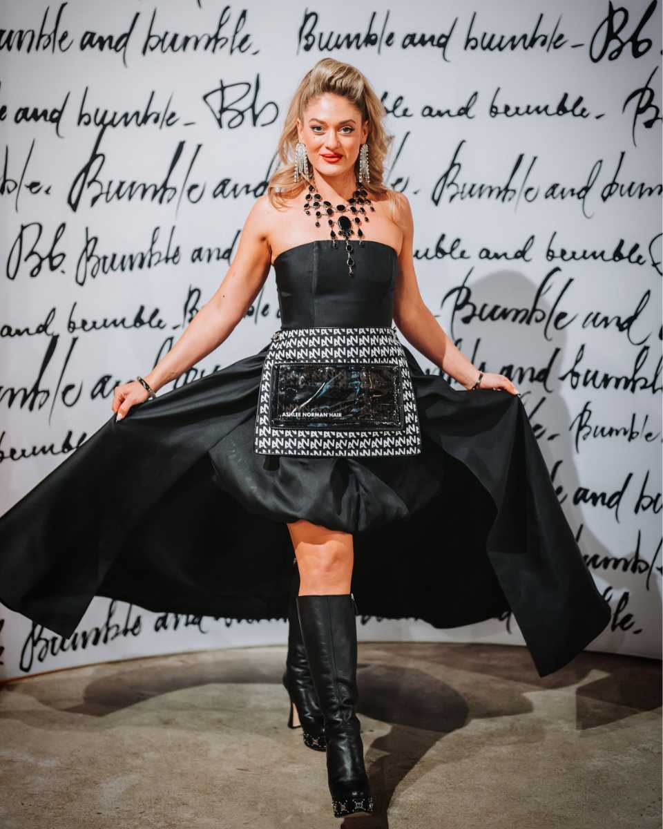 Ashlee Norman poses in front of a Bumble and bumble backdrop wearing a black dress, statement jewelry and boots while holding out the sides of her skirt.