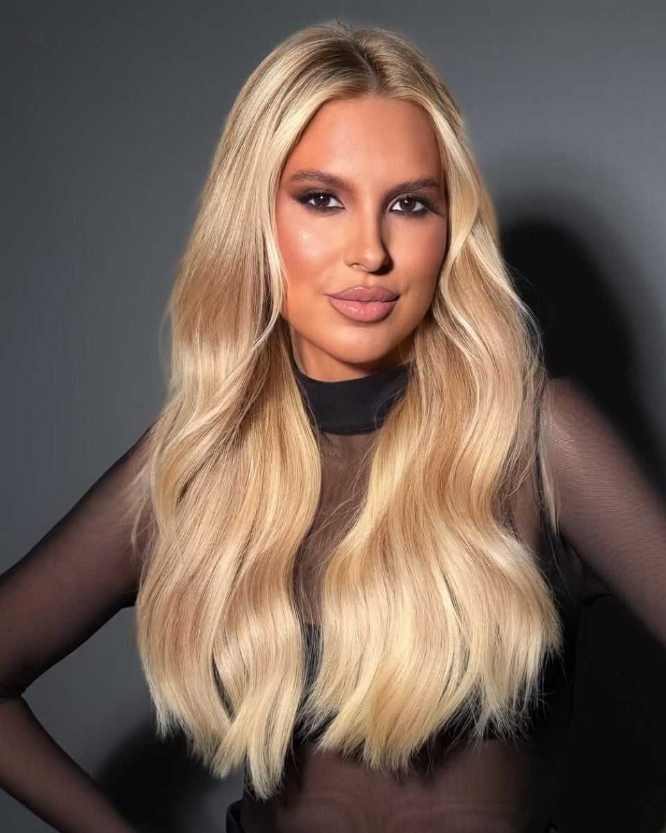 Portrait of long champagne blonde hair with soft beige dimension, a seamless root blend and polished waves styled over a sheer black top.