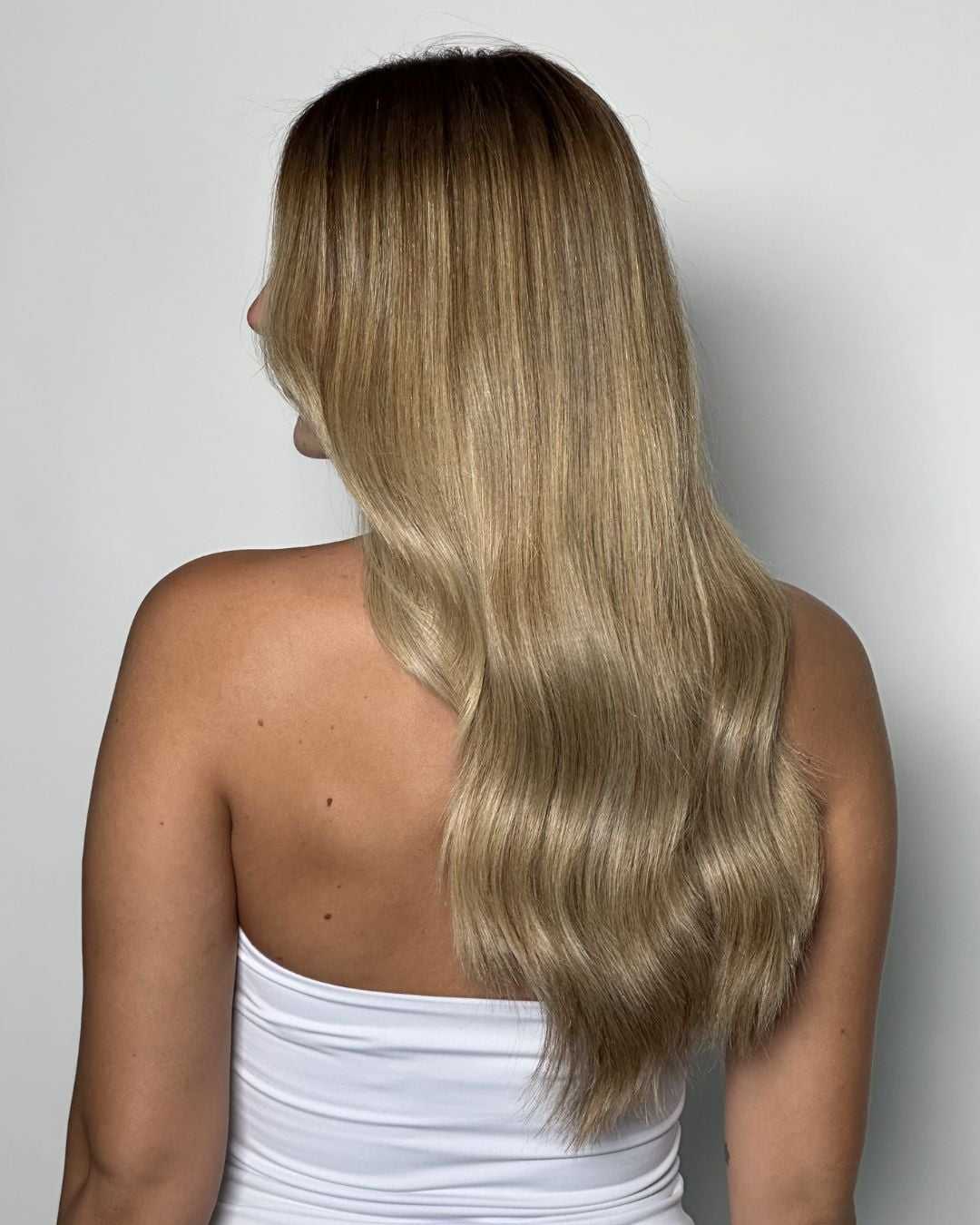 Finished result of long, softly waved blonde hair with a creamy beige-blonde tone. A subtle root shadow melts into luminous blonde lengths, creating a natural, low-maintenance blonde with high shine and dimension.