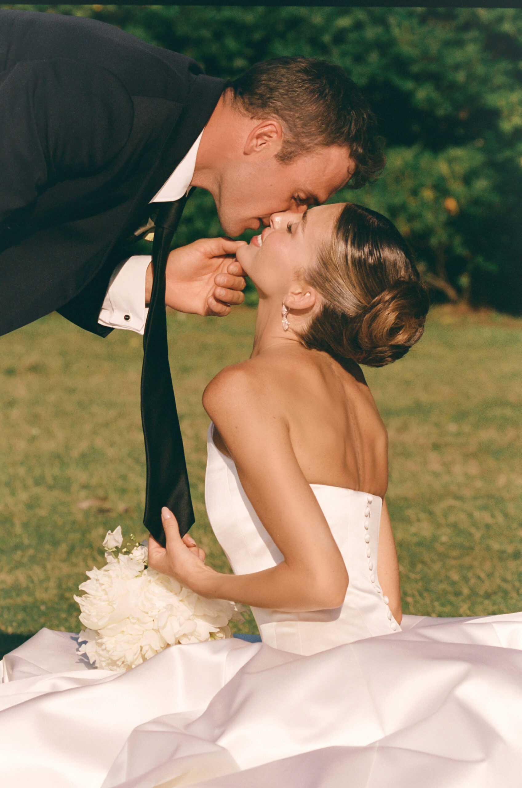 Paige Lorenze shows off her soft brunette highlights in an elegant low bridal bun while sharing a kiss with her husband on their wedding day.