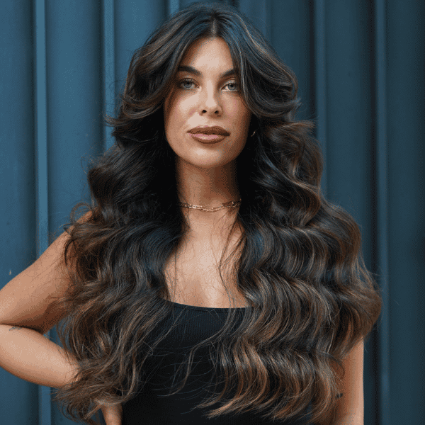 Brunette model with long, dimensional waves and soft face-framing highlights, wearing a black dress, standing against a modern dark blue background