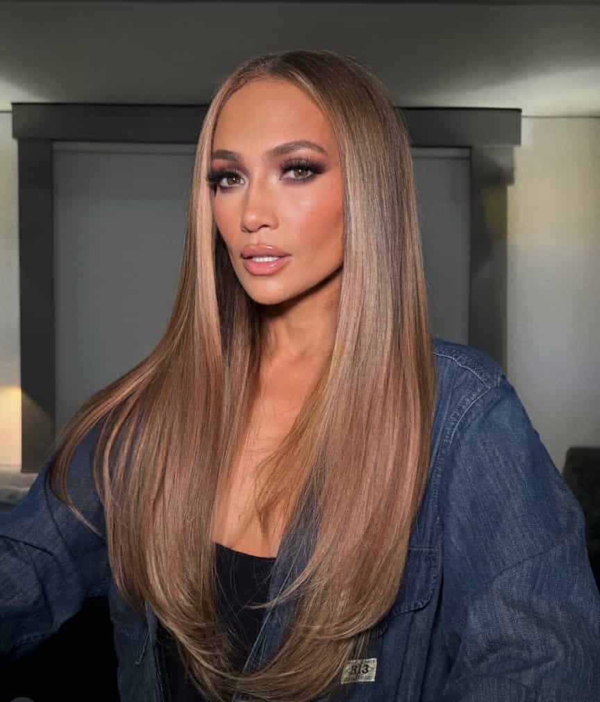 Jennifer Lopez with long, sleek honey-blonde hair styled straight with a center part, wearing a dark denim jacket and neutral glam makeup, sitting in a softly lit indoor setting.
