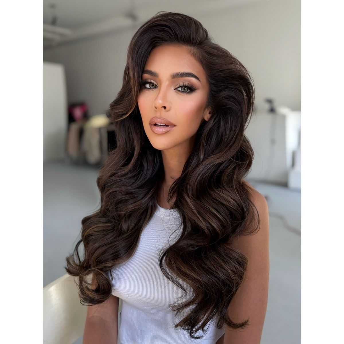 A woman with long, voluminous dark brunette hair styled in glamorous, soft waves. She’s wearing a white sleeveless top and posing in natural light, showcasing glossy, multidimensional color and full-bodied texture.