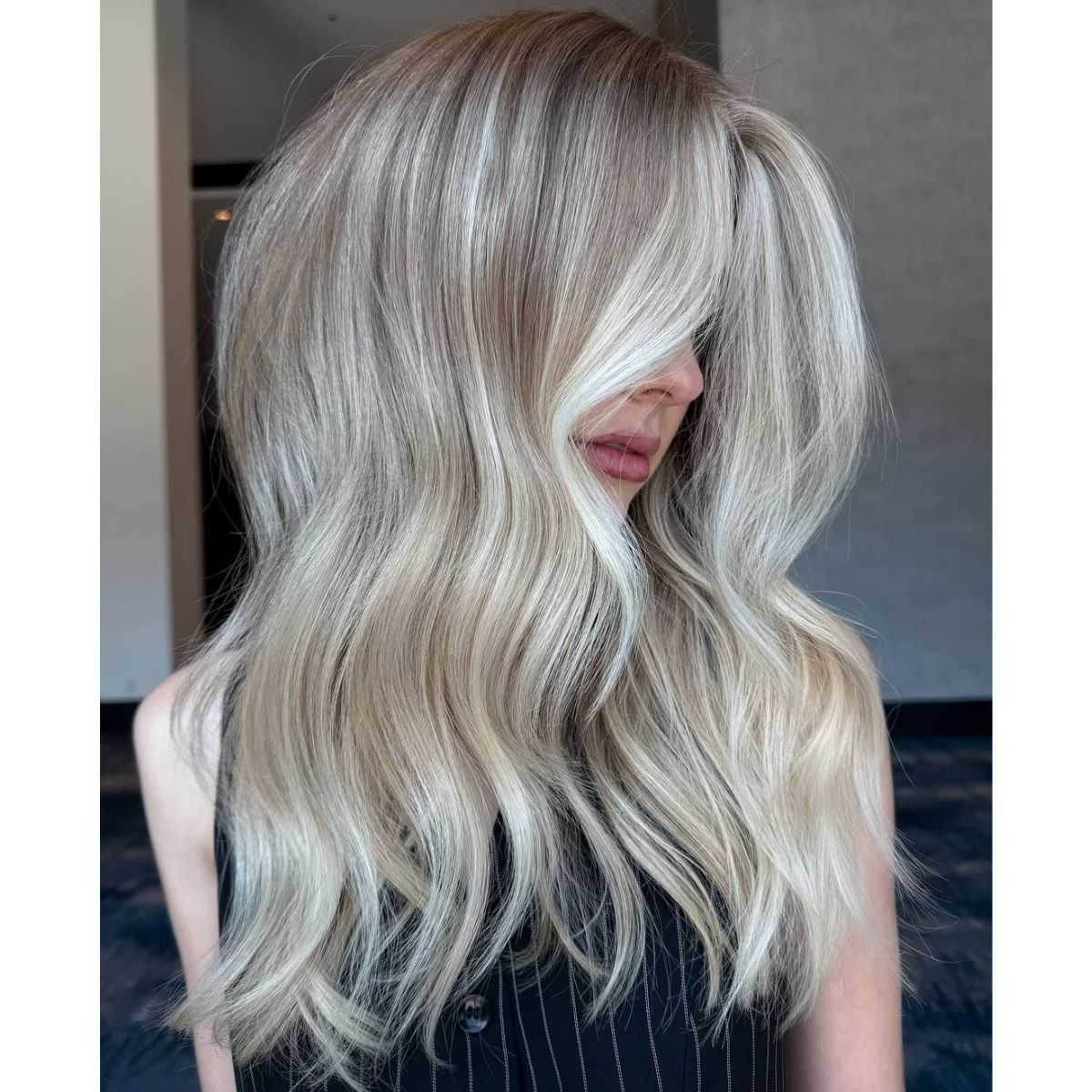 A woman with voluminous, wavy platinum blonde hair and bright, cool-toned highlights, styled with a deep side part and smooth, glossy texture in the same indoor location.