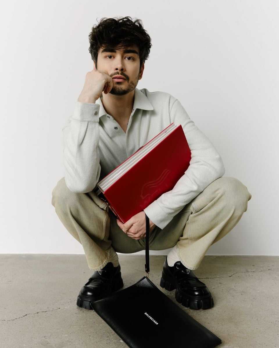 Hairstylist Chris Weber poses holding a red Wella portfolio with a black Balenciaga bag in front of him.