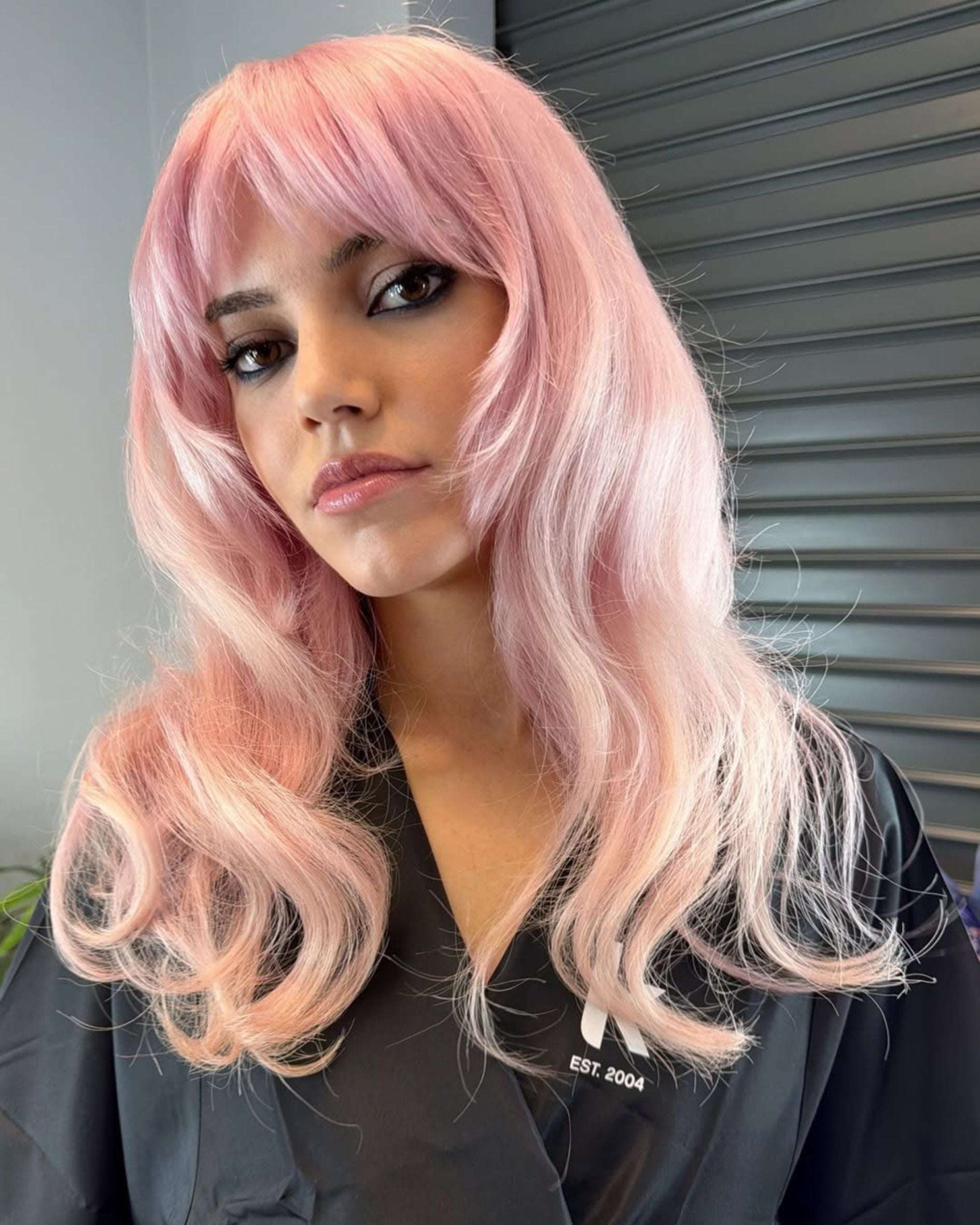 Soft pastel pink hair with long layers and curtain bangs, featuring a sheer, wearable pink tone created on a light blonde base.
