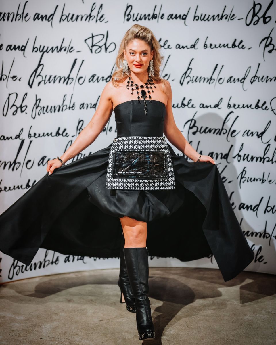 Ashlee Norman poses in front of a Bumble and bumble backdrop wearing a black dress, statement jewelry and boots while holding out the sides of her skirt.