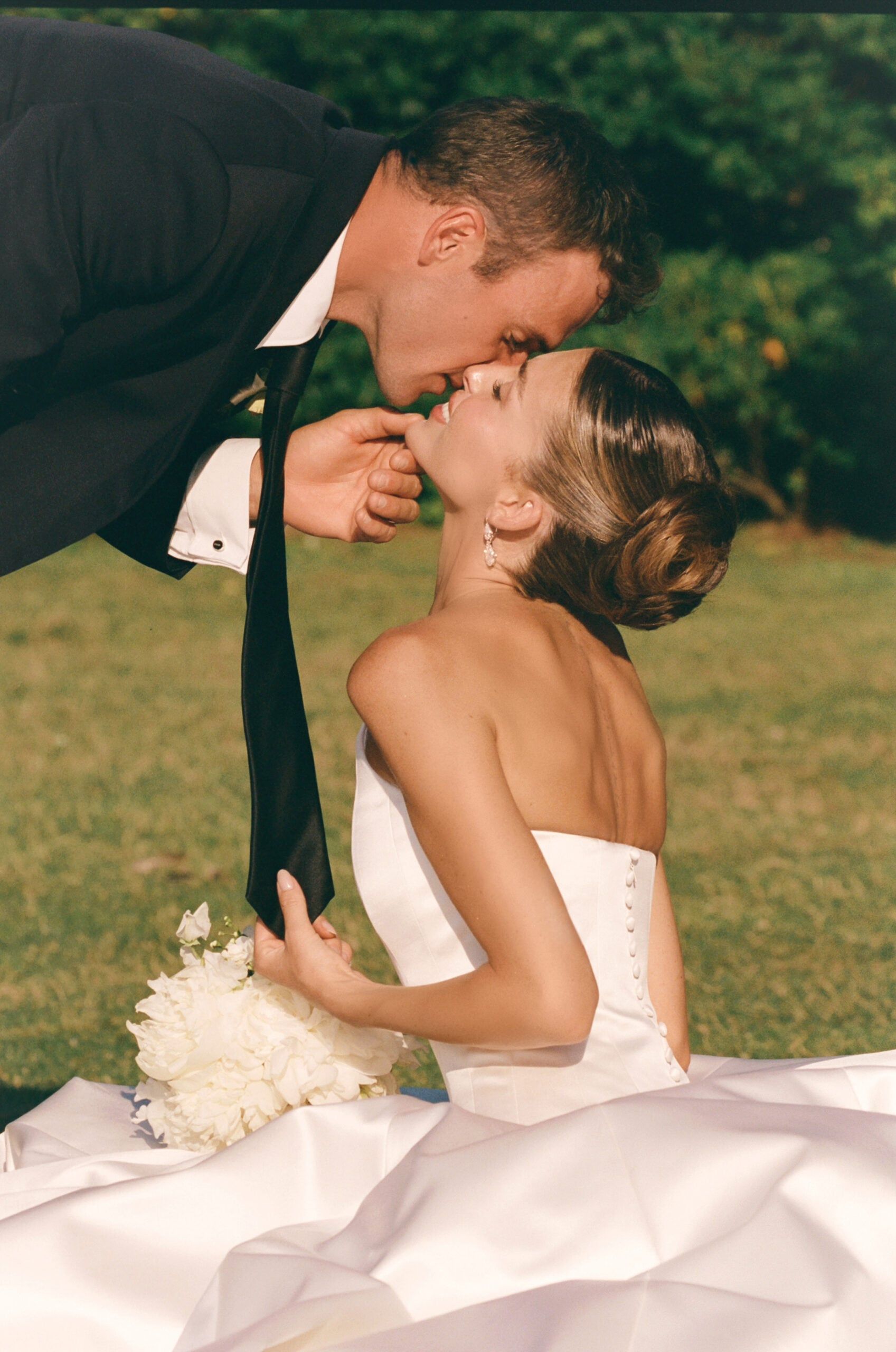 Paige Lorenze shows off her soft brunette highlights in an elegant low bridal bun while sharing a kiss with her husband on their wedding day.