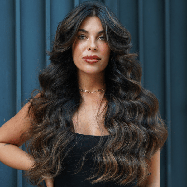 Brunette model with long, dimensional waves and soft face-framing highlights, wearing a black dress, standing against a modern dark blue background
