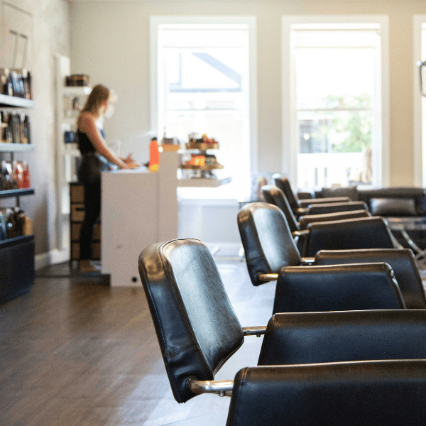 Modern hair salon with black styling chairs and a receptionist at the front desk, featuring bright natural lighting.