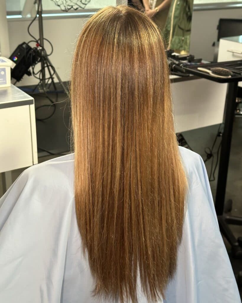 Back view of long, straight blonde hair showcasing a seamless blonde balayage transformation. Fine woven highlights create natural-looking dimension from a darker root into bright beige-blonde mids and ends.